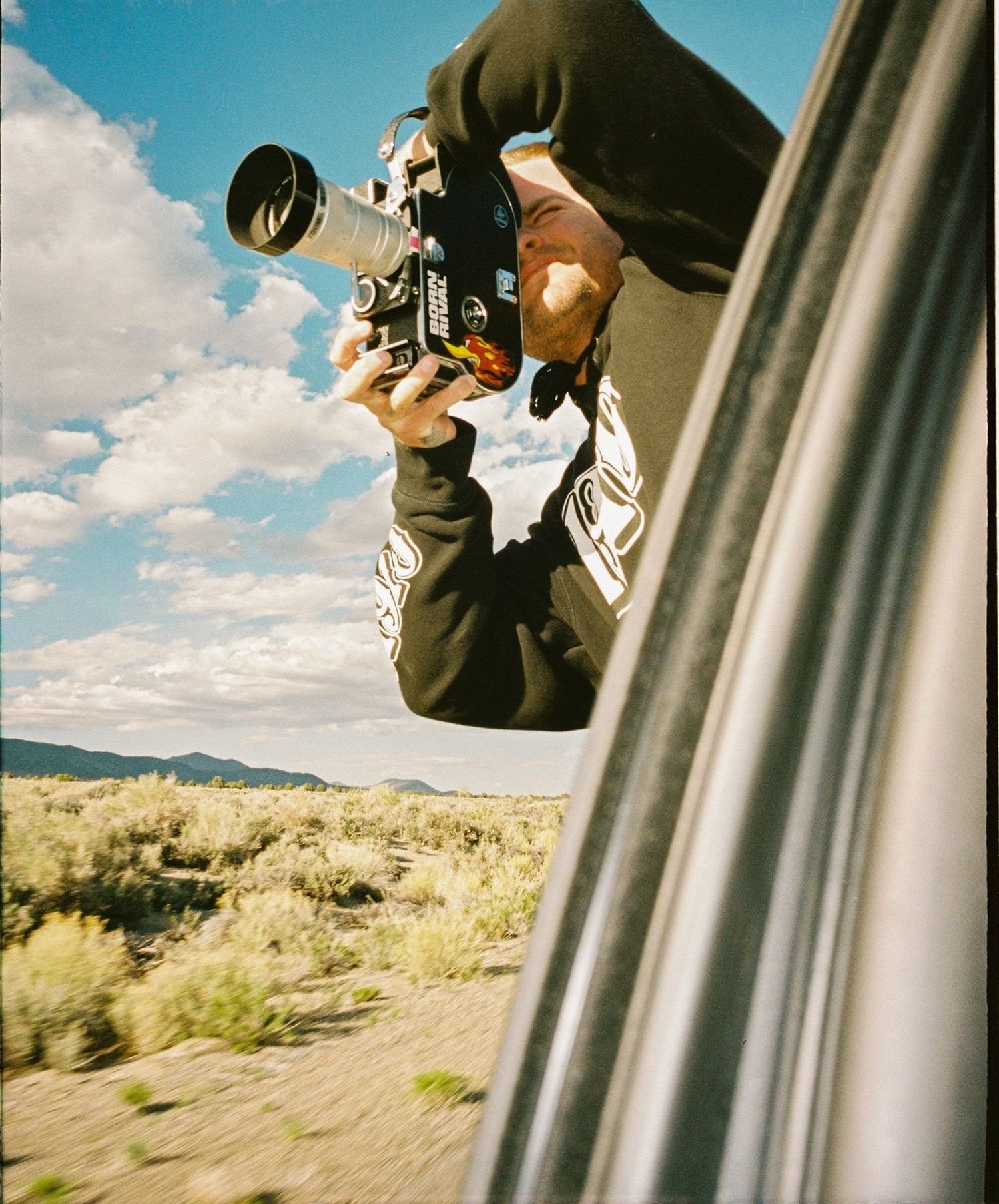 Sam Moody leaning out of a moving car window with a 16mm camera,
             high desert scrub running past below.